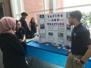 Two students standing by a display board explaining information about wrapping injuries to another student,